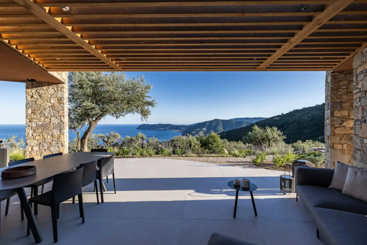 Patio coperto sotto una pergola in legno che incornicia la baia di Alassio e un vecchio ulivo