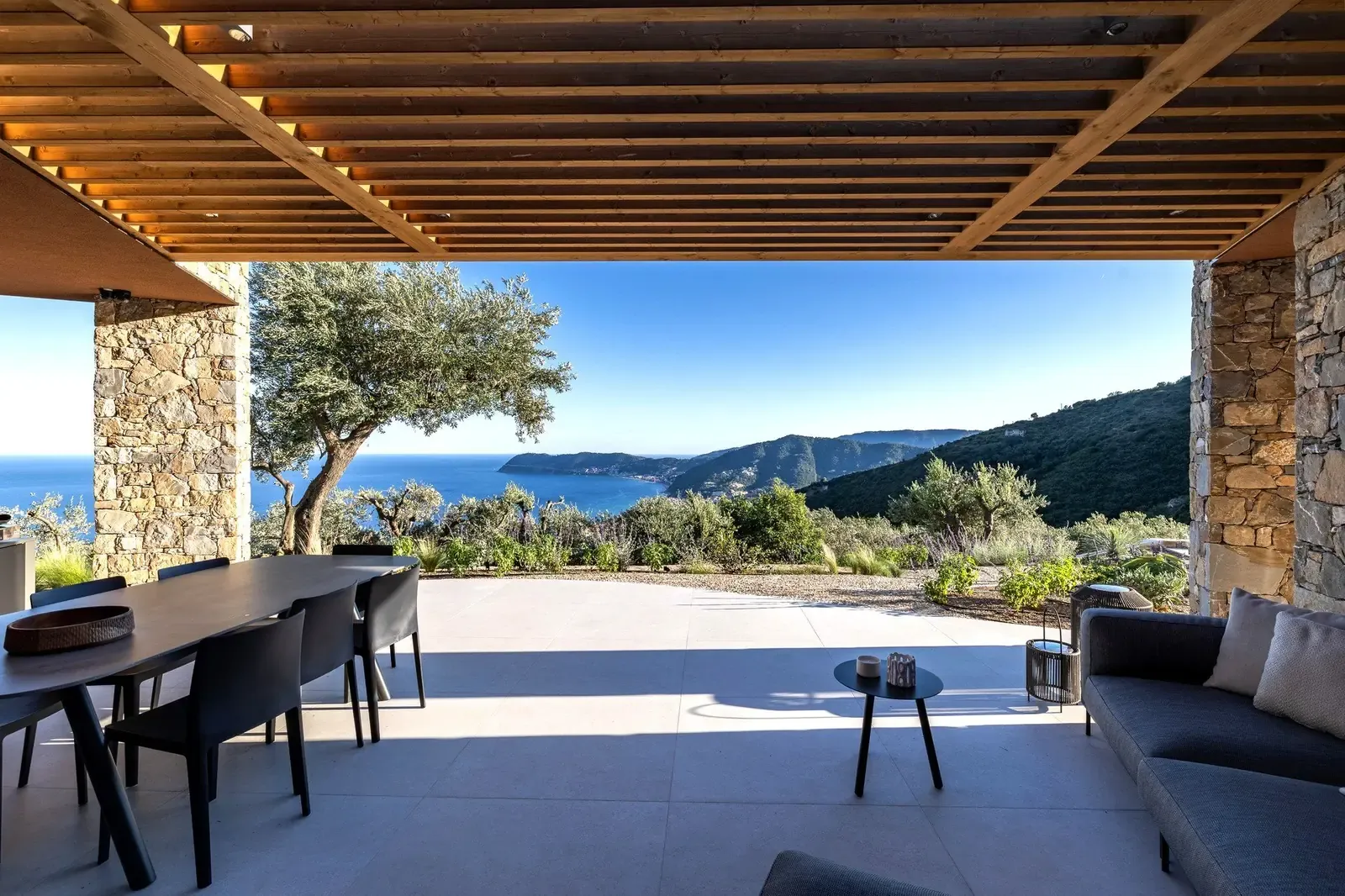 Covered patio under a timber pergola, framing the bay of Alassio and an old olive tree